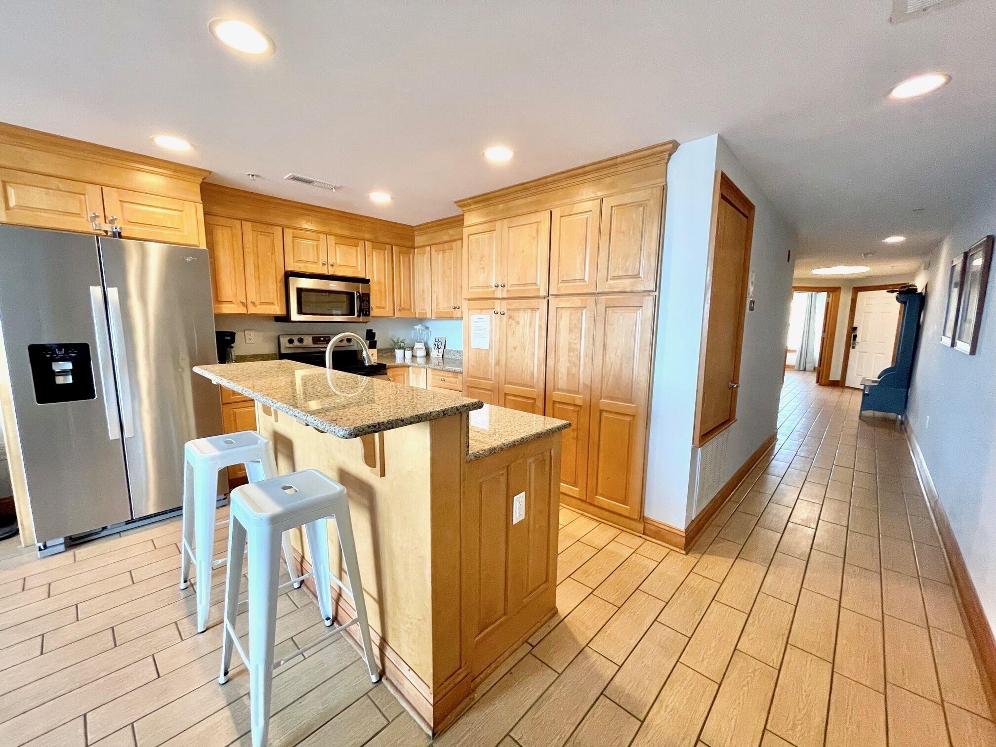 A kitchen with wooden cabinets, a stainless steel refrigerator, a microwave, and a granite countertop island with stools.