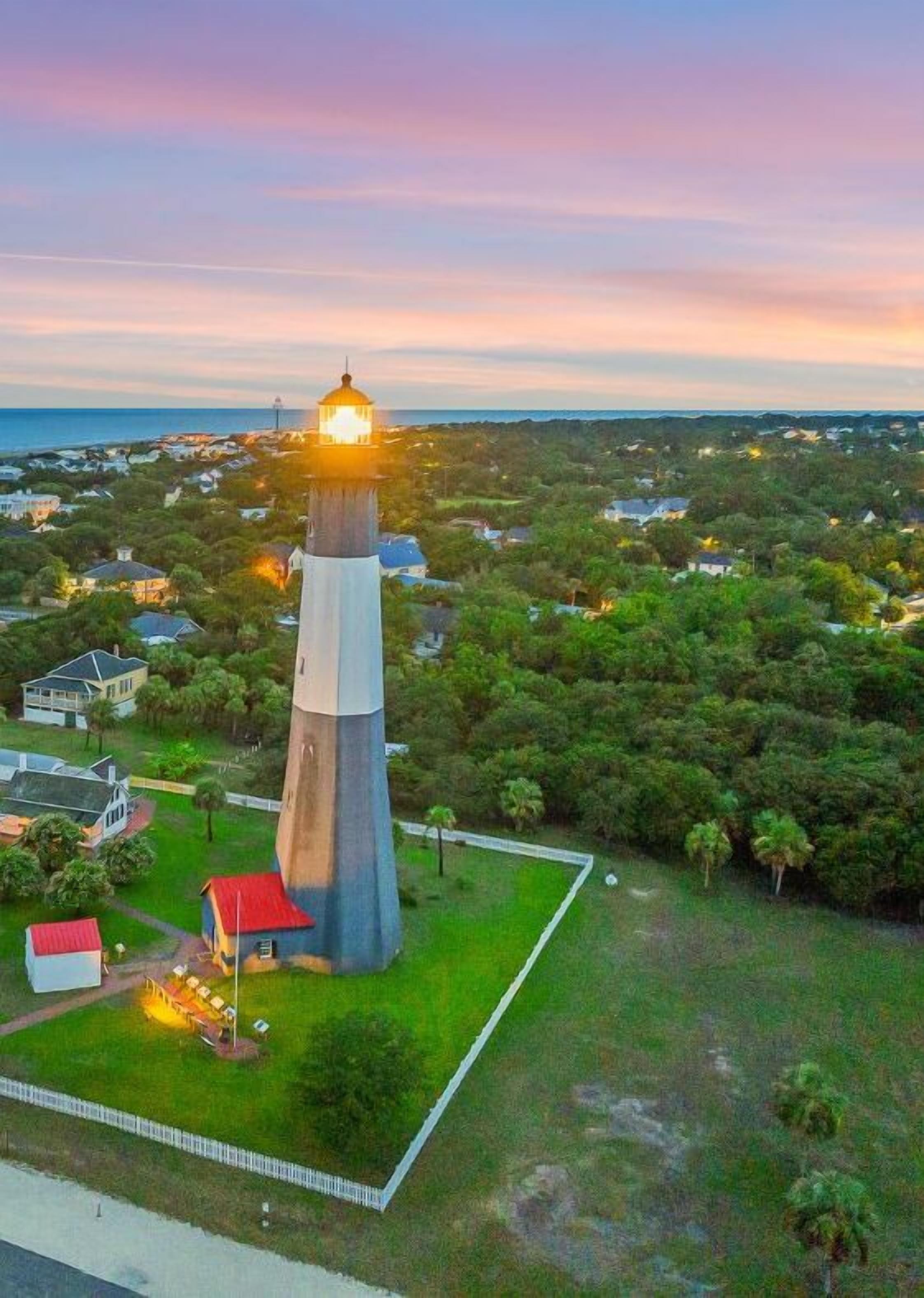 A lighthouse with a light at the top, surrounded by a grassy area and a few buildings nearby.