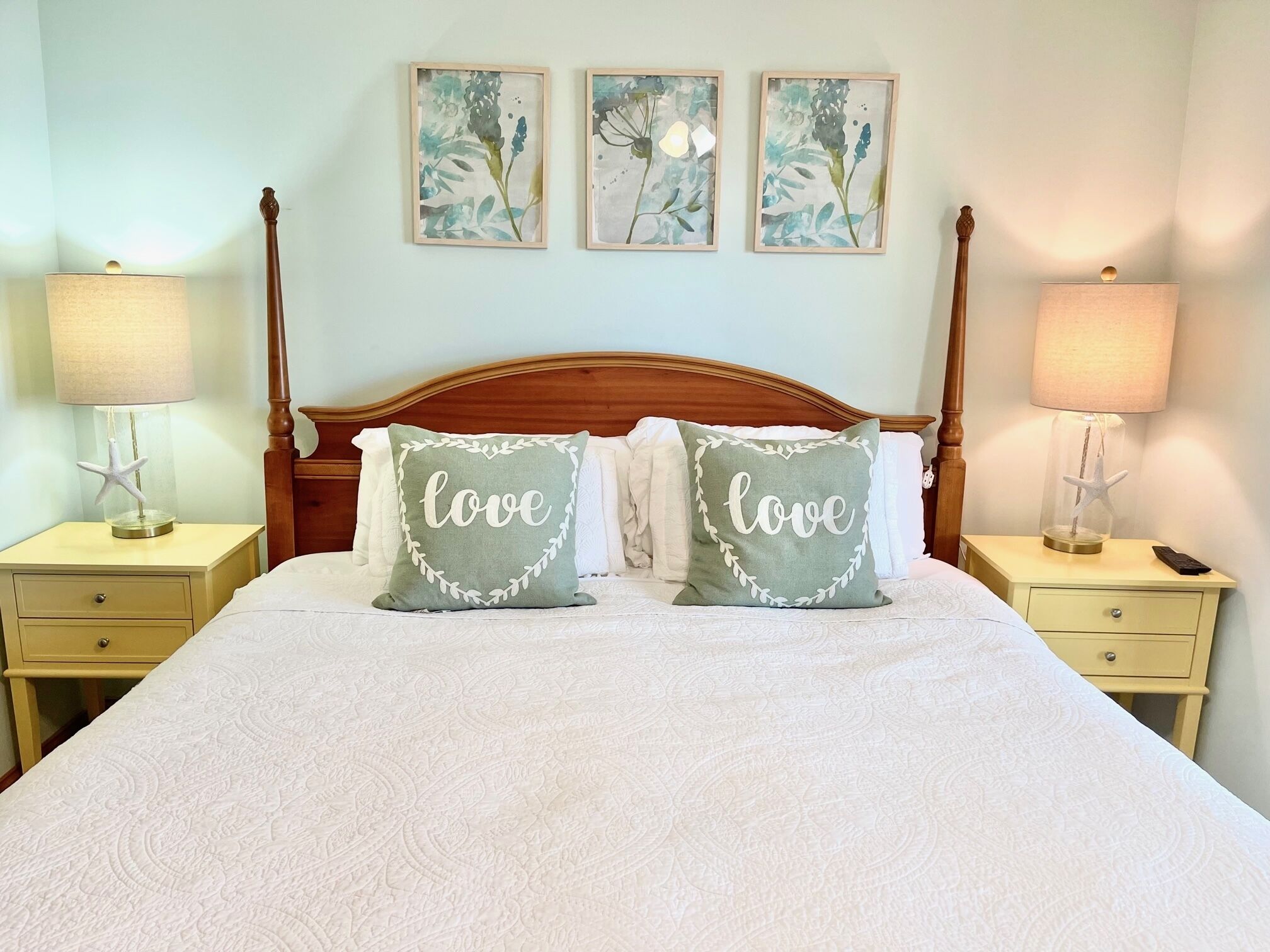 Un letto rifatto con cura, con due cuscini, ognuno con la scritta "love", affiancato da due comodini con lampade e un tavolino con un telecomando.