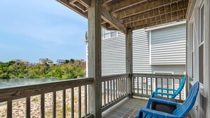 A wooden deck with a railing overlooking a beach, two blue chairs, and a view of houses in the distance. - Outer Banks Bliss: 3-bedroom Cottage in Golf Community, Beach Club Access (Nags Head)