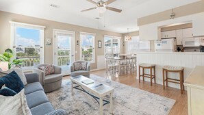 A spacious living room with a large window, a ceiling fan, and a dining area with a table and chairs. - Outer Banks Bliss: 3-bedroom Cottage in Golf Community, Beach Club Access (Nags Head)