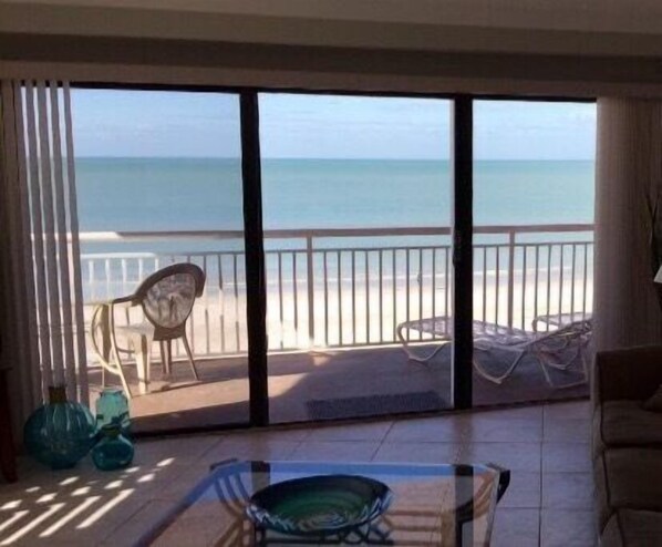 Une pièce dotée d’une grande porte-fenêtre coulissante donnant sur un balcon avec vue sur la plage. Le balcon est agrémenté de deux chaises et la pièce comprend une table avec un plateau en verre.