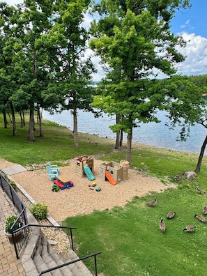A lakeside park with a playground, a small bridge, and a grassy area with geese.