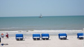 Una playa con varias sillas azules dispuestas en la arena, un velero a lo lejos y un cielo azul despejado.
