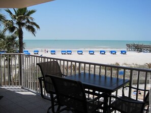A balcony with a table and chairs overlooking a beach with blue umbrellas and a pier.