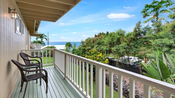 A wooden deck with wicker chairs, a white railing, and a view of the ocean and tropical vegetation.