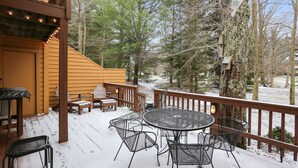 A wooden deck with outdoor furniture, a snow-covered ground, and a view of trees.