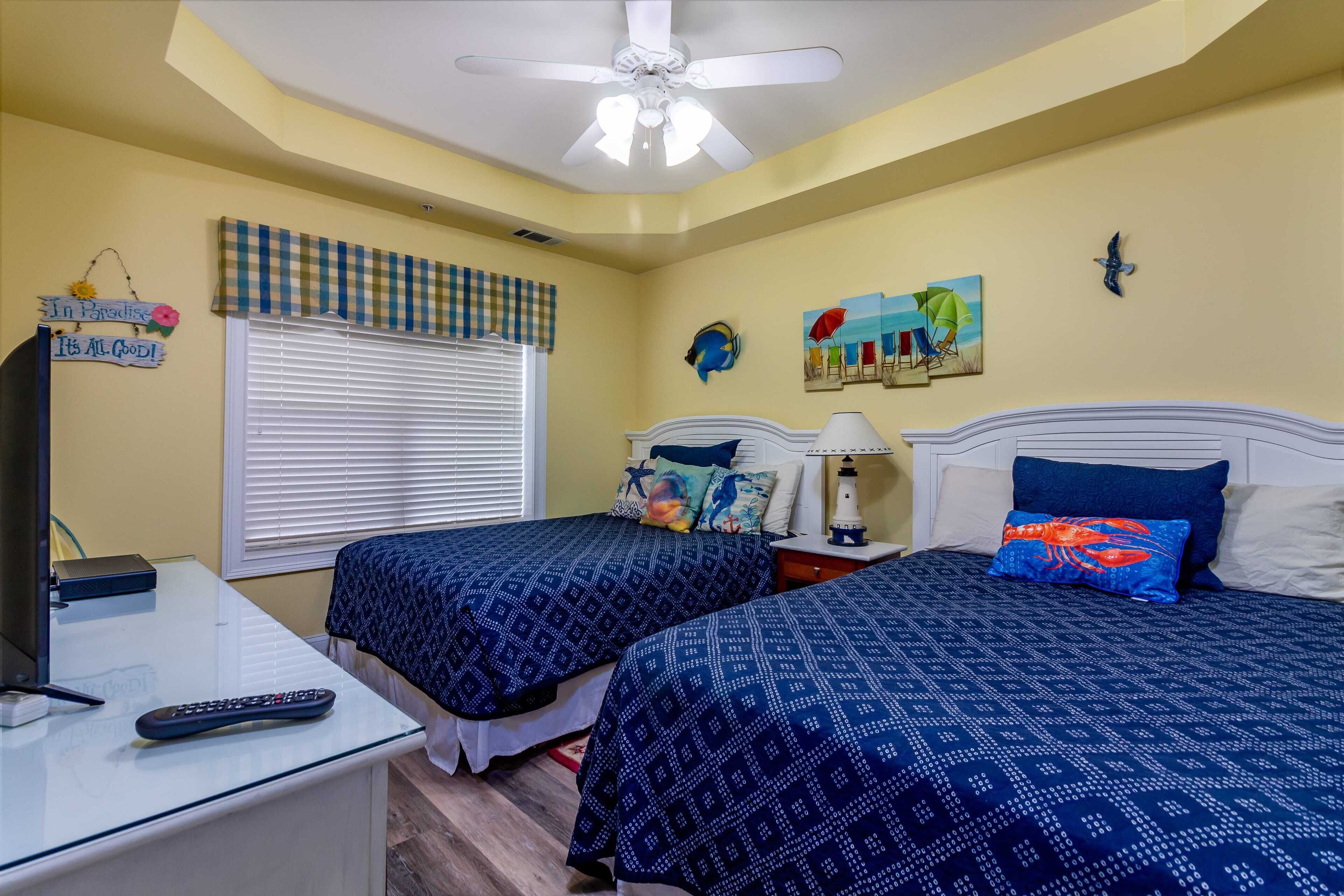 A bedroom with two beds, a ceiling fan, a window with blinds, a nightstand with a lamp, and wall decorations including a painting and a sign.