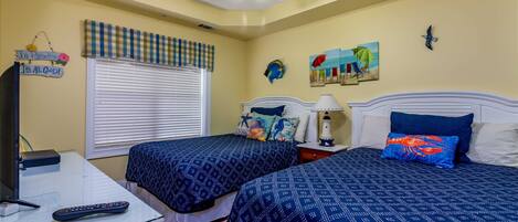 A bedroom with two beds, a ceiling fan, a window with blinds, a nightstand with a lamp, and wall decorations including a painting and a sign.