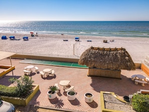 Un patio frente a la playa con mobiliario exterior, una estructura de techo de paja y vista al océano.
