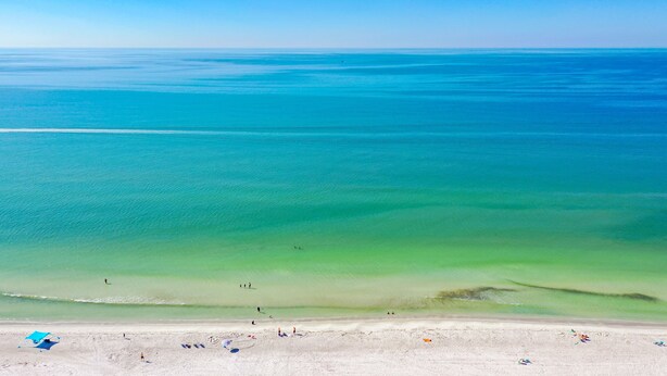 A beach with clear turquoise water, a sandy shore, and a few umbrellas. - Beach Palms 507 -Gulf front luxurious Condo - Spectacular Views! (Indian Shores)