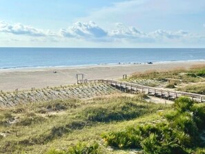 Ein Strand mit einem hölzernen Promenadenweg, grasbewachsenen Dünen und dem Meer im Hintergrund.