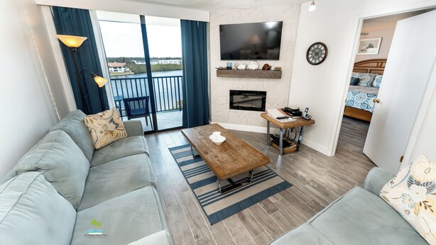 A modern living room with a sofa, coffee table, and TV. There is a fireplace, a balcony with curtains, and a view of a body of water. - Quiet Waters Condominium 5B (Indian Shores)
