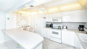 A modern kitchen with white cabinets, a central island, a microwave, a stove, and a coffee maker. - Quiet Waters Condominium 5B (Indian Shores)