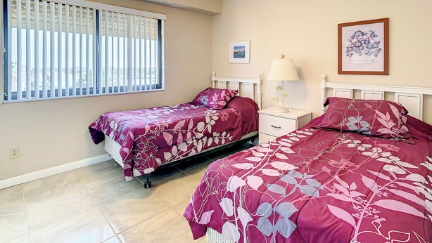 A bedroom with two beds, a ceiling fan, a window with blinds, a bedside table with a lamp, and a framed picture on the wall.