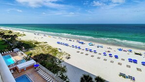 A beach with rows of sun loungers and umbrellas, a pool area, and a view of the ocean.