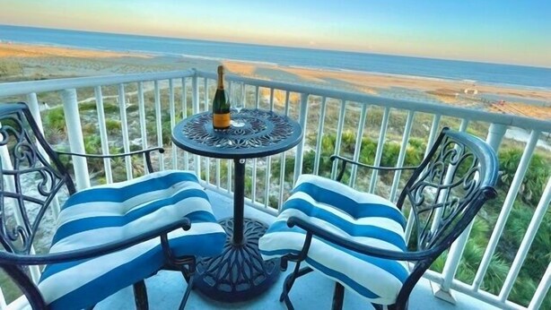 A balcony with two striped chairs and a table, overlooking a beach. - Summer & July 4th Savings at Beachside Colony Tybee's ONLY Oceanfront Resort 3/2 (Tybee Island)