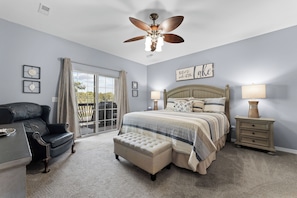 Une chambre avec un grand lit, un ventilateur de plafond, une vue à travers une porte-fenêtre coulissante et un fauteuil en cuir noir.