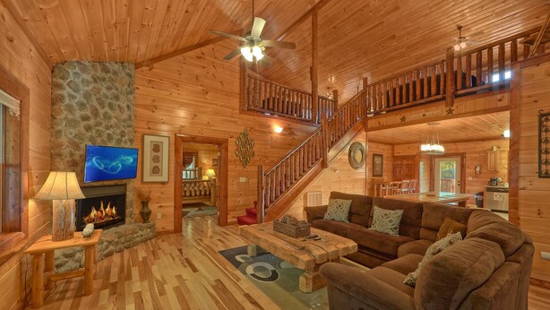 A spacious log cabin interior with a fireplace, a large sofa, a dining area, and a staircase leading to an upper level. - Live, Laugh, Love Pool Lodge: Indoor Pool, Theater (Cosby)