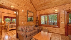 A cozy cabin interior with a wooden floor, a sofa, a coffee table, and a large window offering a view of the outdoors. - Live, Laugh, Love Pool Lodge: Indoor Pool, Theater (Cosby)