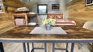 A rustic cabin interior with a wooden table, a metal bucket with flowers, a bed with a red and white checkered blanket, and a window with curtains. - W-W: Pavilion and Glamping Village (Monticello)