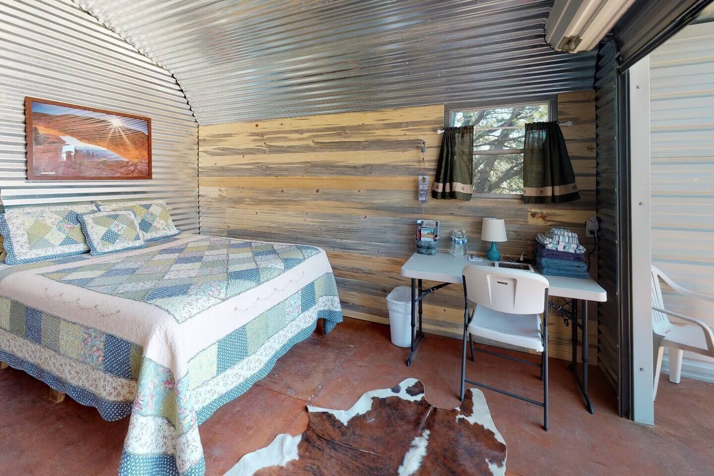 A bedroom with a bed, a desk, a chair, a cowhide rug, a painting, and a window.