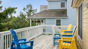 Una terraza de madera con sillas Adirondack azules y amarillas, una barandilla blanca y un edificio de color azul claro con tejado de tejas.