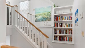 A staircase with white railings and wooden handrails, leading to a landing with a bookshelf and a painting of a beach scene. - Southern Reach: Soundside Golf Villa w/ Beach Club Access and Sunset Views (Nags Head)