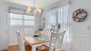 A dining room with a wooden table, white chairs, a chandelier, and a window with blinds. - Southern Reach: Soundside Golf Villa w/ Beach Club Access and Sunset Views (Nags Head)