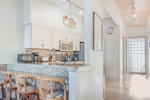 A kitchen with white cabinets, a granite countertop, a toaster, a microwave, and a coffee maker. There is a wicker chair at the counter and a clock on the wall.