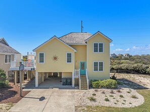 A two-story yellow house with a front porch, a basketball hoop, and a small garden area.