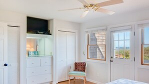 Ein Schlafzimmer mit einem Bett, einem Deckenventilator, einem Fernseher, einem Sessel und einem Fenster mit Aussicht.