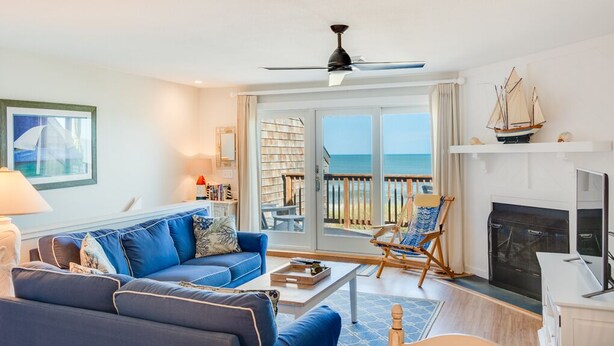 Ein Wohnzimmer mit blauem Sofa, Couchtisch, Kamin, Fernseher und einem Balkon mit Meerblick.