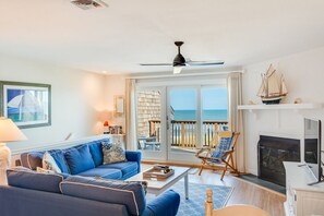 A living room with a blue sofa, a coffee table, a fireplace, a television, and a balcony with a view of the ocean. - Whispering Sands #6: Oceanfront Condo, Shared pool (Nags Head)