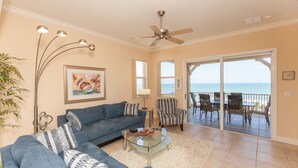A living room with a blue sofa, a glass coffee table, and a view of the ocean through a sliding glass door. - Cinnamon Beach 741 - Direct Oceanfront Corner Unit! New Living Room! (Palm Coast)