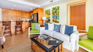 A modern kitchen with wooden cabinets, a dining area with wicker chairs, a white sofa with colorful cushions, and a glass coffee table. - Late Summer Savings • Beachside Colony • Tybee’s ONLY Oceanfront Resort• 2BR/2BA (Tybee Island)