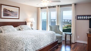 A bedroom with a large bed, a ceiling fan, a chair, a table, and a view of the sea. - Sound, Mind and Bodie: Condo, Shared Pool (Manteo)