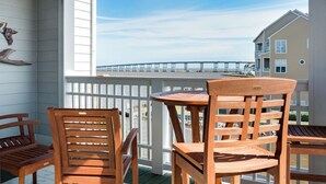 A wooden deck with two chairs and a table, overlooking a bridge and beach. - Sound, Mind and Bodie: Condo, Shared Pool (Manteo)