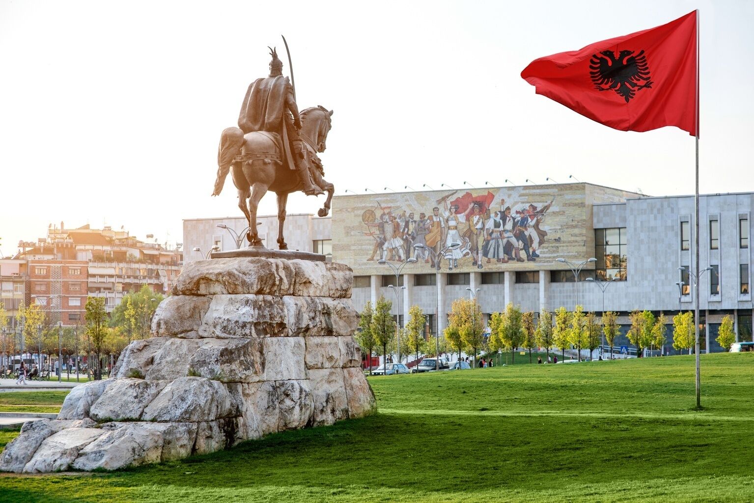 A statue of a mounted figure on a rock pedestal, a flagpole with a flag, and a building with a mural in the background.
