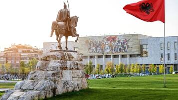 A statue of a mounted figure on a rock pedestal, a flagpole with a flag, and a building with a mural in the background.