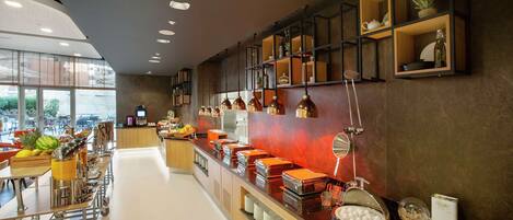 Un moderno bufé de desayuno con una variedad de alimentos y bebidas, una estación de café y una zona de estar con mesas y sillas.