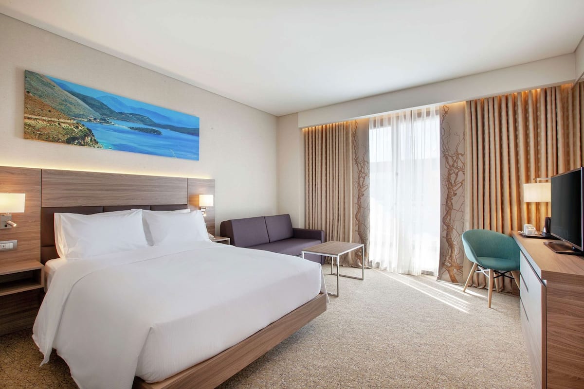 A hotel room with a large bed, a sofa, a desk with a TV, and a painting of a coastal landscape.