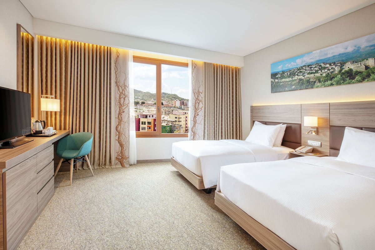 A hotel room with two beds, a desk, a chair, and a large window with a city view.