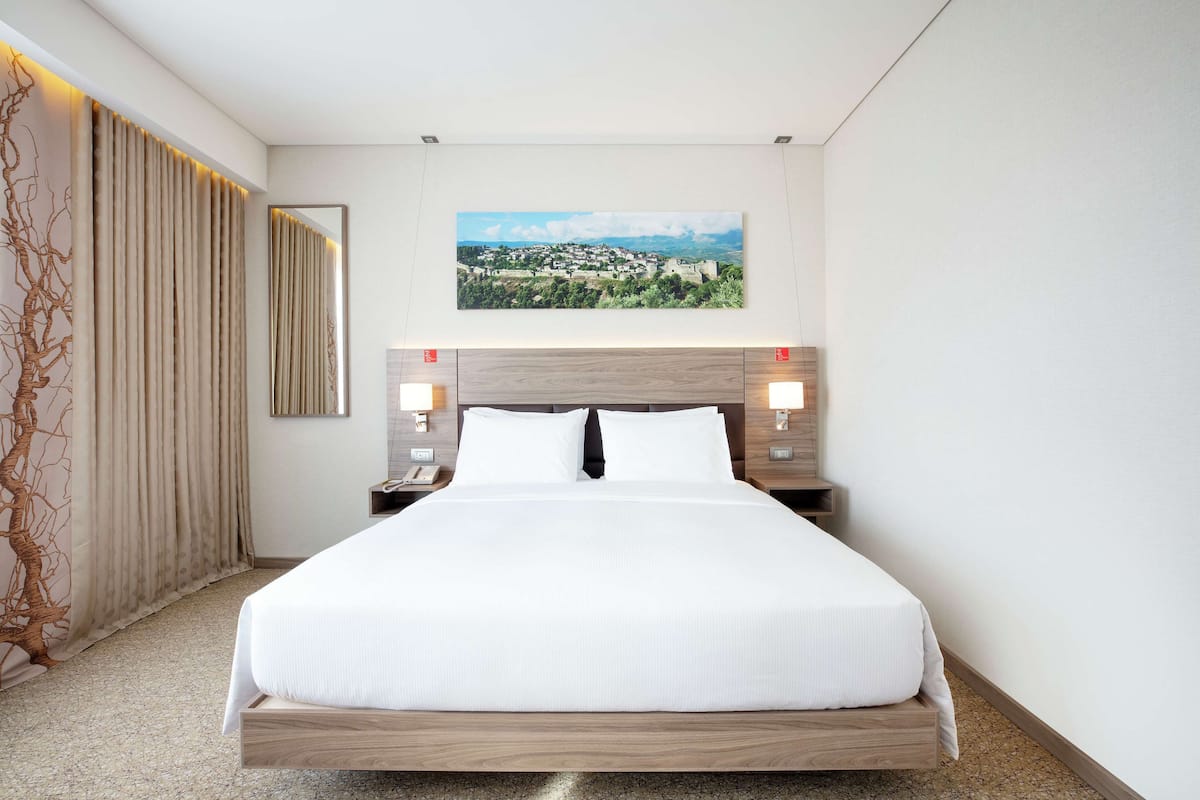 A hotel room with a large bed, a view of a town, and a decorative wall piece.