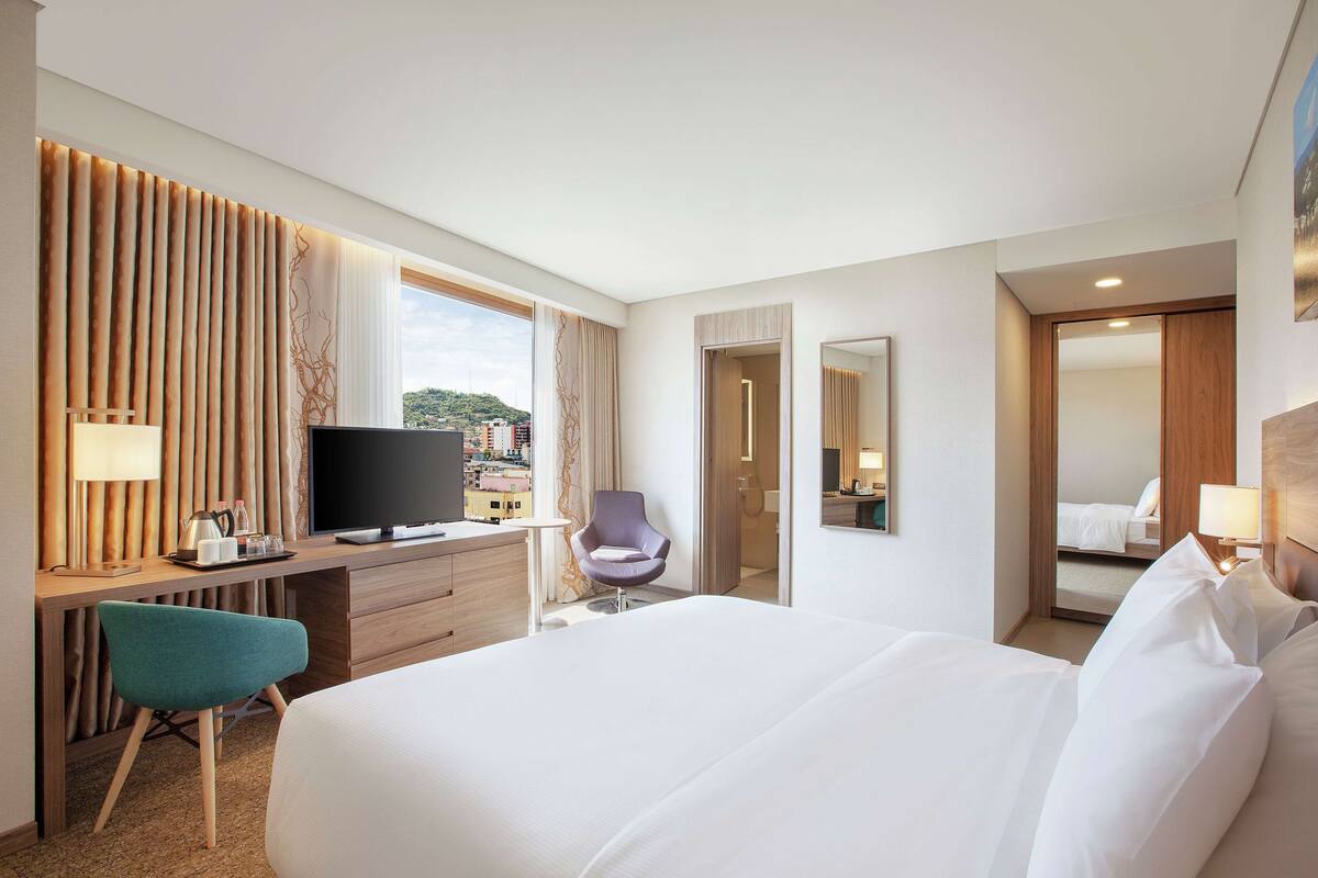 A hotel room with a large bed, a desk with a TV, a chair, and a window with curtains.