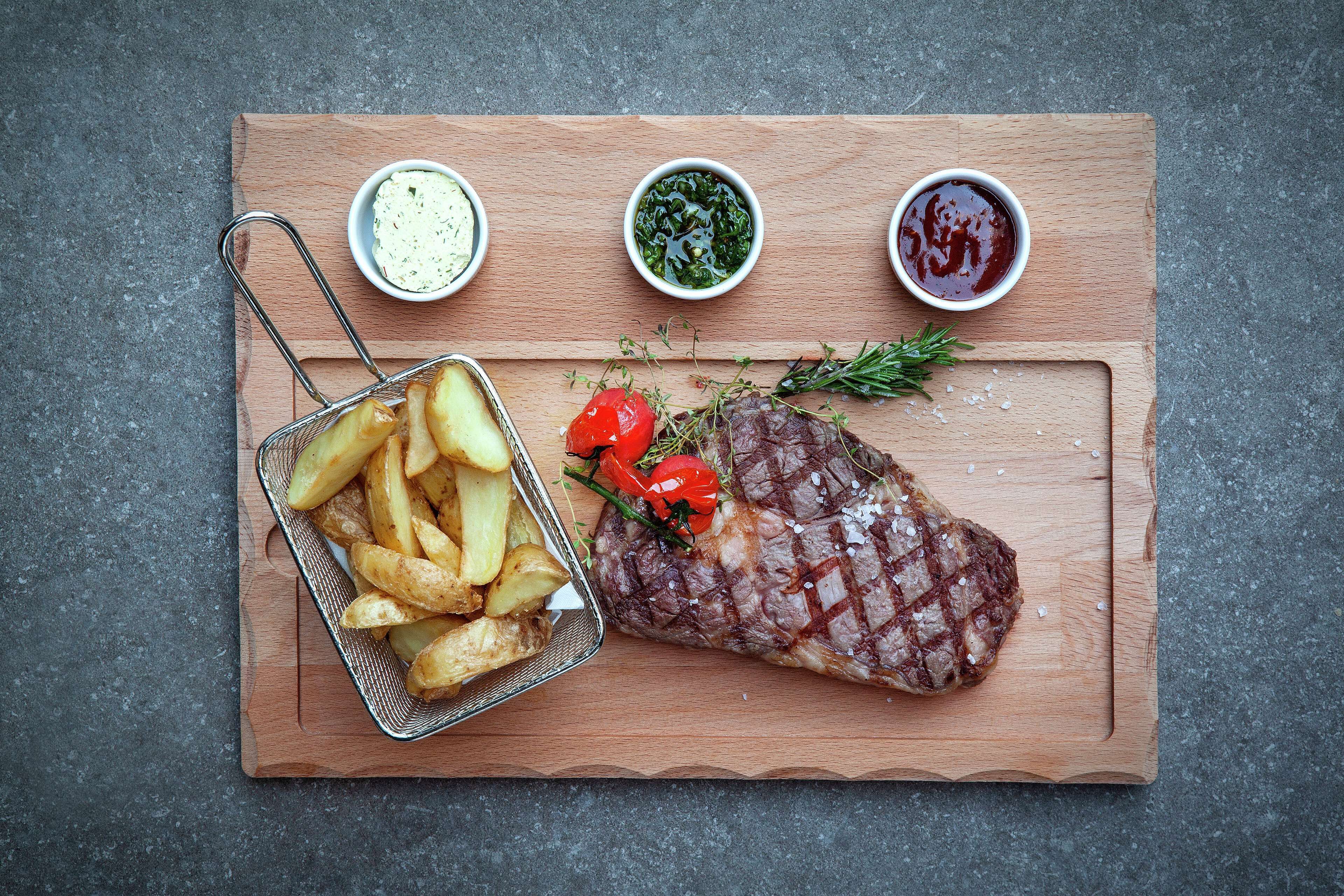 A wooden board with a grilled steak, a basket of fries, and three small bowls of sauces.