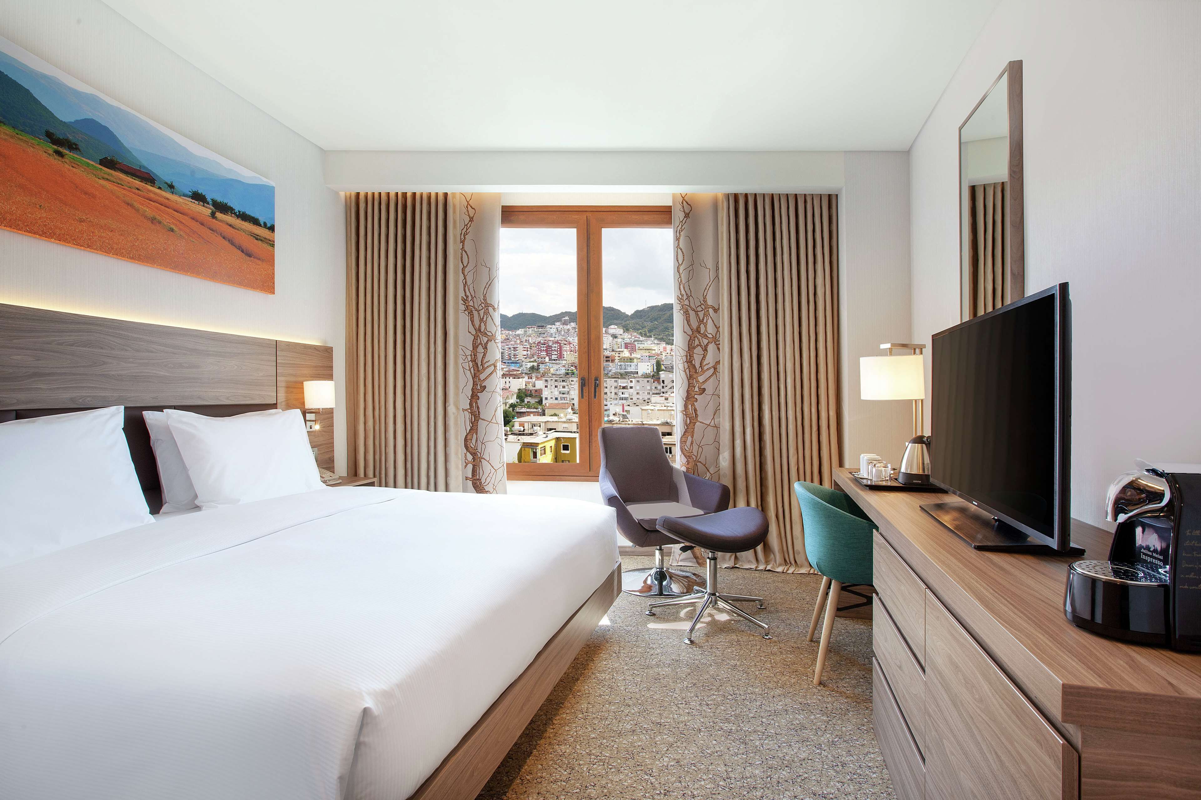 A hotel room with a large bed, a desk with a TV, a chair, and a view of a cityscape through the window.
