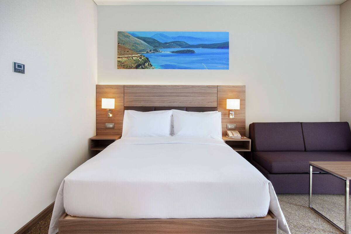 A hotel room with a large bed, a wooden headboard, two bedside tables with lamps, a dark-colored sofa, and a painting of a coastal landscape on the wall.