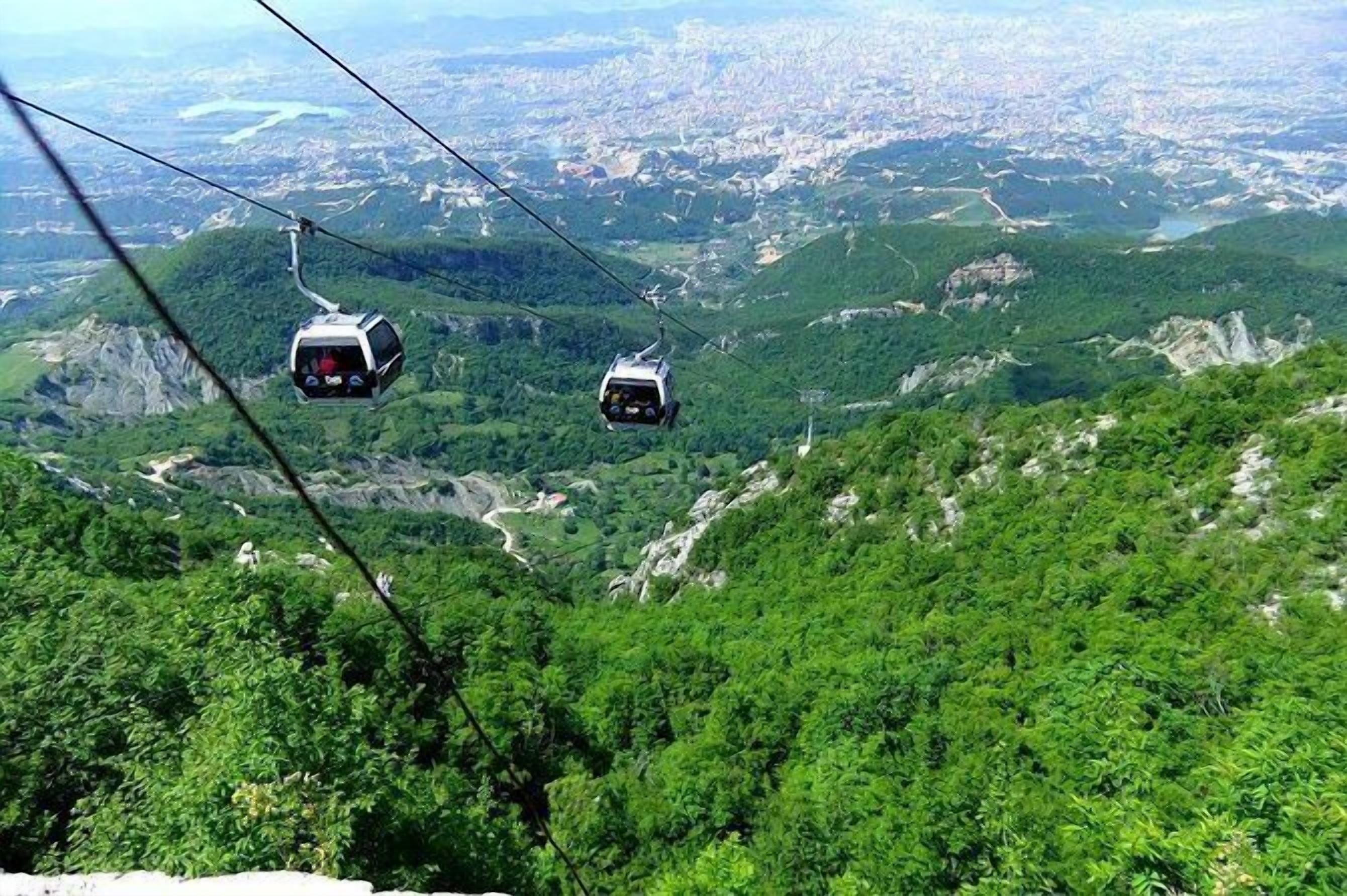 Eine Gondelbahn in einer gebirgigen Landschaft mit üppiger Vegetation und einer ferngesiedelten Stadtansicht.