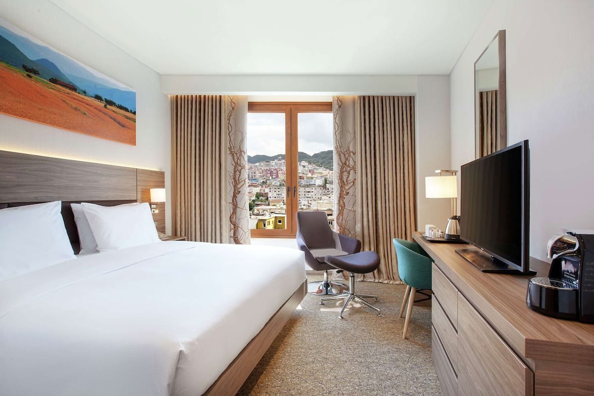 A hotel room with a bed, a desk with a TV, a chair, and a view of a cityscape through a large window.
